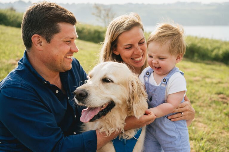 Parents with a young child and a golden retriever, sitting together and smiling during a family photo session near the sea in Torbay