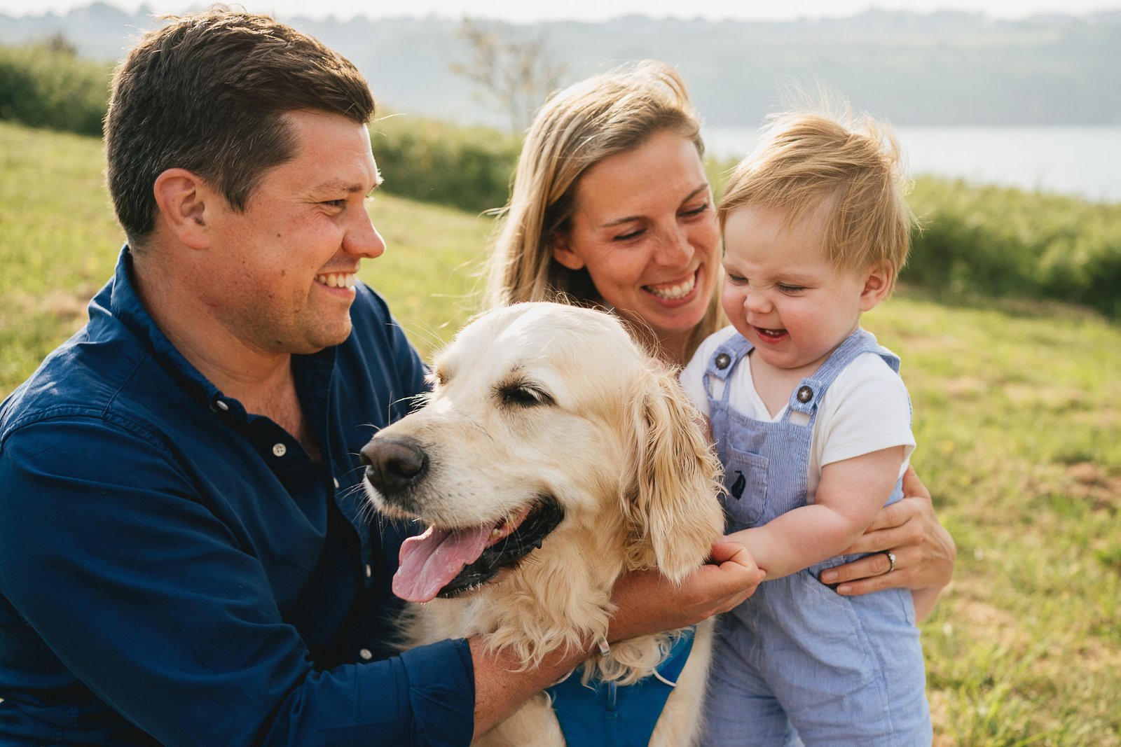 Parents with a young child and a golden retriever, sitting together and smiling during a family photo session near the sea in Torbay